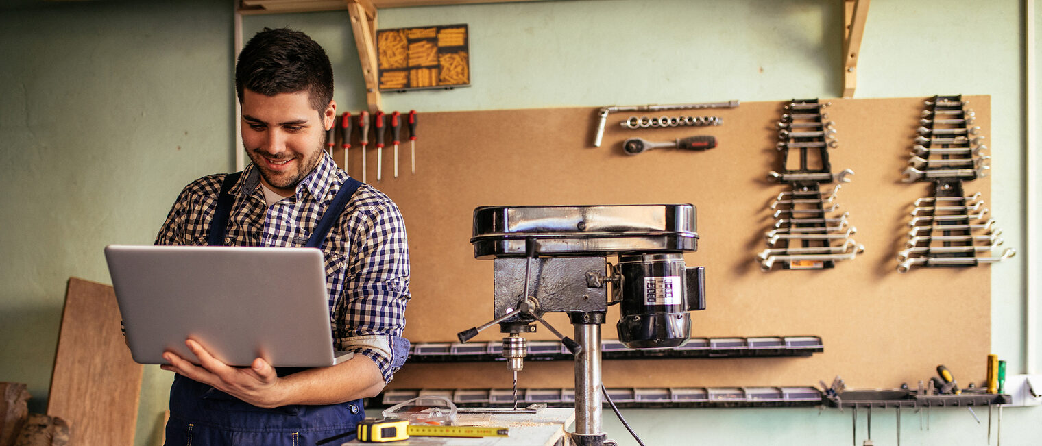 Marketing im Handwerk Ein Handwerker mit einem Laptop in seiner Werkstatt. Im Hintergrund hängen verschiedene Werkzeuge.