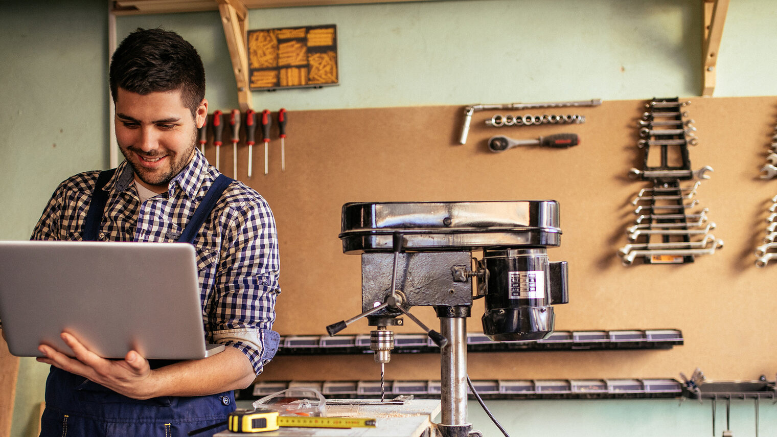 Ein Handwerker mit einem Laptop in seiner Werkstatt. Im Hintergrund hängen verschiedene Werkzeuge.