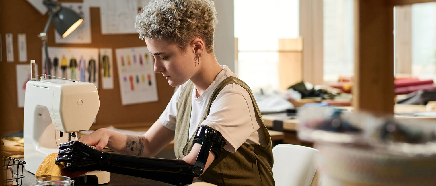 Inklusion im Handwerk Eine Schneiderin mit Armprothese näht ein Kleidungsstück mit einer Nähmaschine.