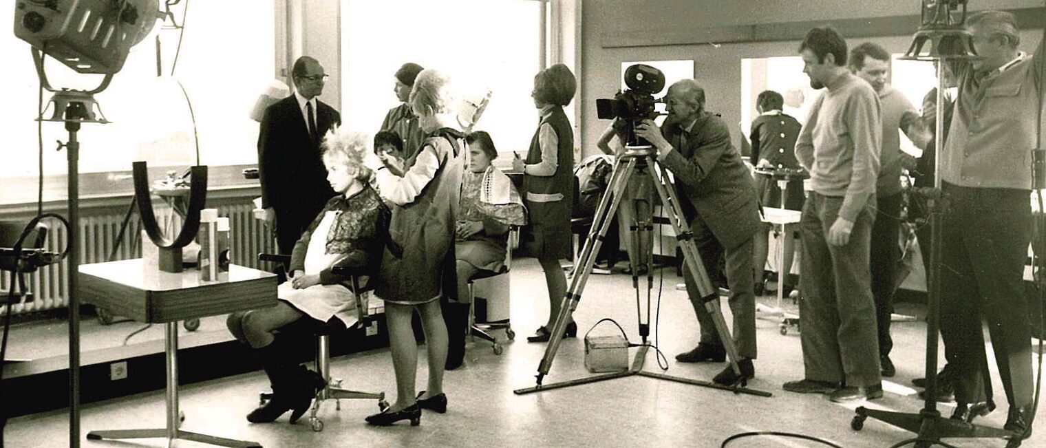 Ein Filmset, aufgebaut im bbz Arnsberg. Zu sehen sind Scheinwerfer, Mitarbeiter und ein Kameramann, der eine Friseurin filmt.