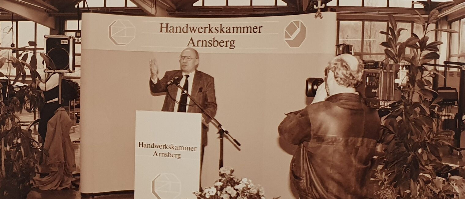 Ein Mann hält eine Rede an einem Rednerpult, das in einer KFZ-Werkstatt aufgebaut ist. Er wird von einem Kameramann gefilmt.