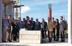 Eine Gruppe von etwa 15 Personen steht auf einem Baugelände hinter einer kleinen Mauer. Ein Mann hält eine Rede.