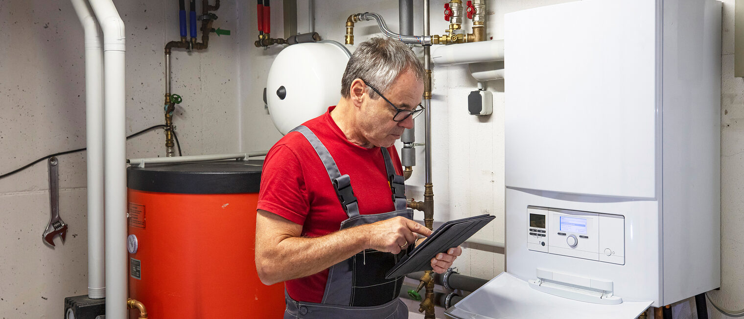 SHK Ein Handwerker in einem Heizungskeller. Er schaut auf ein Tablet, während er neben der Heizung steht.