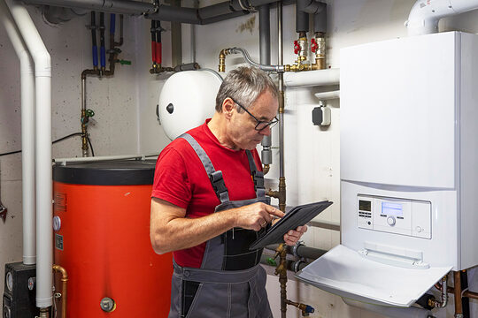 Ein Handwerker in einem Heizungskeller. Er schaut auf ein Tablet, während er neben der Heizung steht.