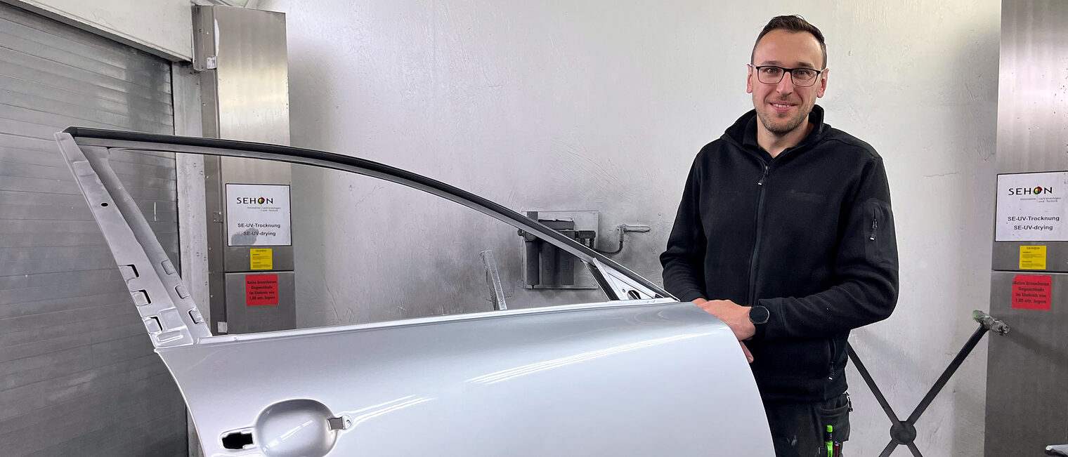 Fahrzeuglackierermeister Simon Schmeinta steht in einer Werkstatt und hält eine silberne Autotür fest.