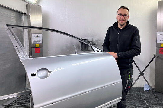 Fahrzeuglackierermeister Simon Schmeinta steht in einer Werkstatt und hält eine silberne Autotür fest.