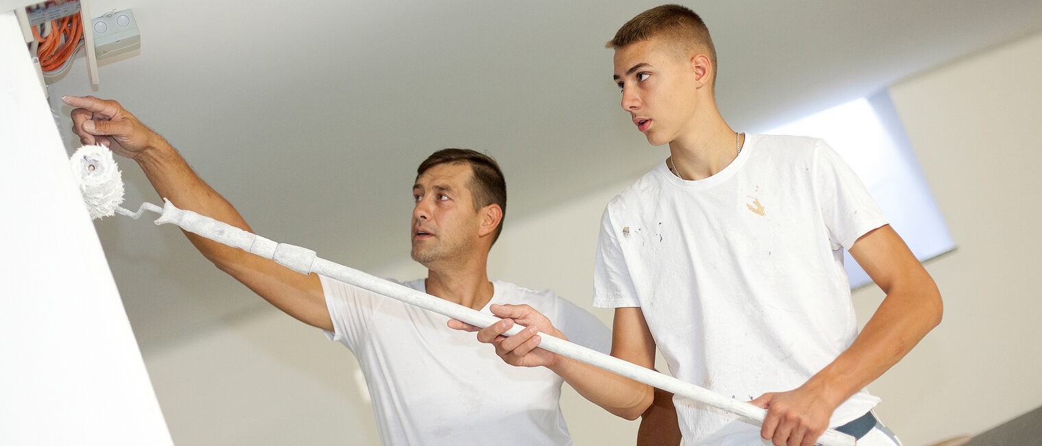 Ausbildung Ein Auszubildender im Bereich Maler und Lackierer streicht eine Wand mit weißer Farbe. Ein Ausbilder erklärt ihm dabei etwas.