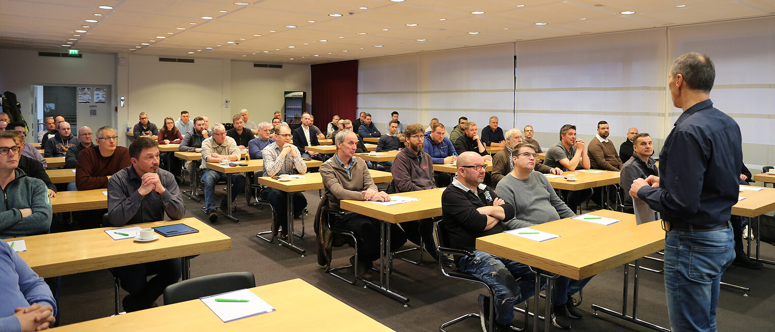 Voller Kursraum am bbz Arnsberg. Ein Dozent steht vorne und trägt vor, die Kursteilnehmer sitzen an den Tischen und hören zu.