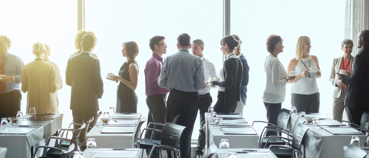 Symbolbild eines Netzwerktreffens. Mehrere Menschengruppen stehen in einem Konferenzraum und sprechen miteinander.