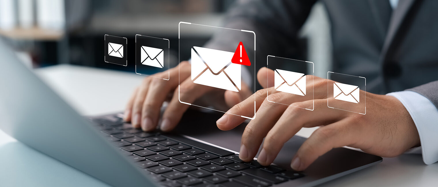 Ein E-Mail-Symbol mit einem roten Warndreieck. Im Hintergrund ist ein Mann zu sehen, der mit einem Laptop arbeitet.