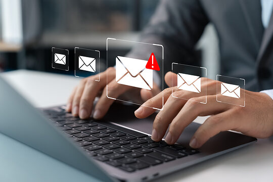 Ein E-Mail-Symbol mit einem roten Warndreieck. Im Hintergrund ist ein Mann zu sehen, der mit einem Laptop arbeitet.
