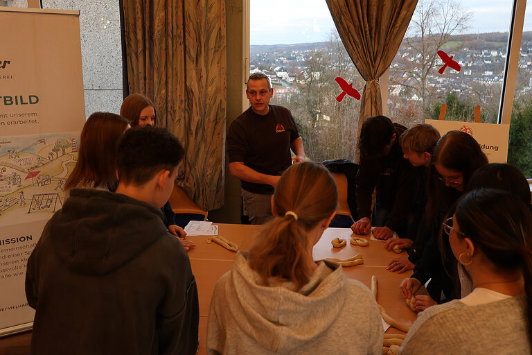 Mehrere Schüler stehen im Kreis um einen Tisch, auf dem Teigrollen liegen. Ein Vertreter der Bäckerei Vielhaber erklärt etwas