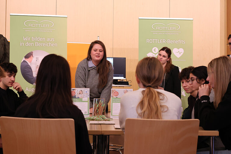 Eine Gruppe von Schülern sitzt an einem Tisch. Zwei Mitarbeiterin eines Augenoptikerbetriebes erklären etwas.