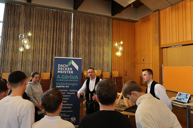 Zwei Handwerker vom Dachdeckerbetrieb Görlich erklären einer Gruppe von Schülern etwas über das Dachdeckerhandwerk.