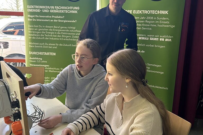 Zwei Mädchen schauen auf einen Computer-Bildschirm. Ein Mann schaut ihnen über die Schulter. Im Hintergrund stehen Werbebanne