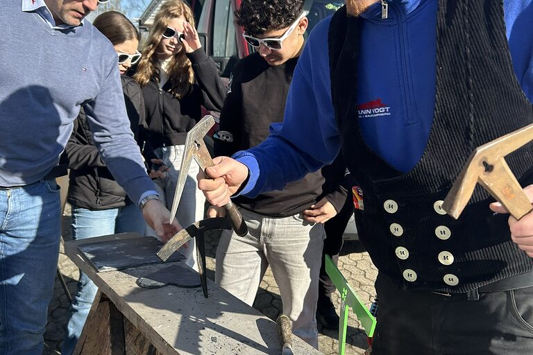 Ein Mann in Handwerkskluft steht an einer Werkbank im Freien. Im Hintergrund stehen ein weiterer Mann und eine Schülergruppe.
