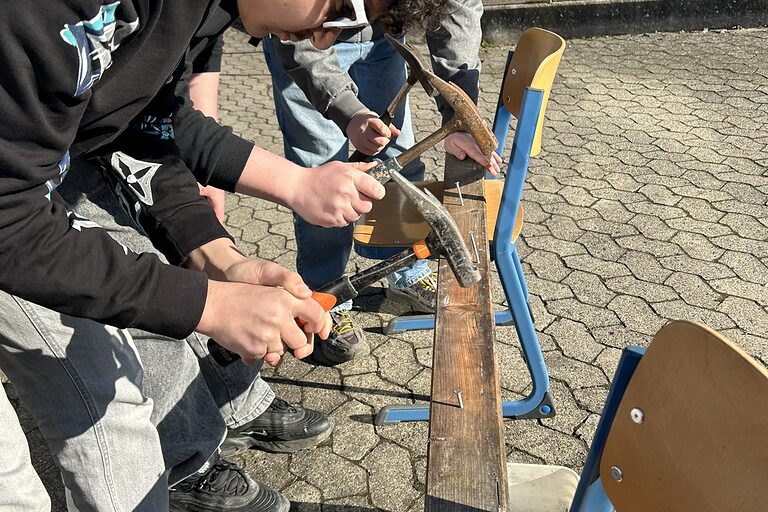Zwei Schüler im Freien nageln mit einem Hammer einen Nagel in ein Holzbrett.