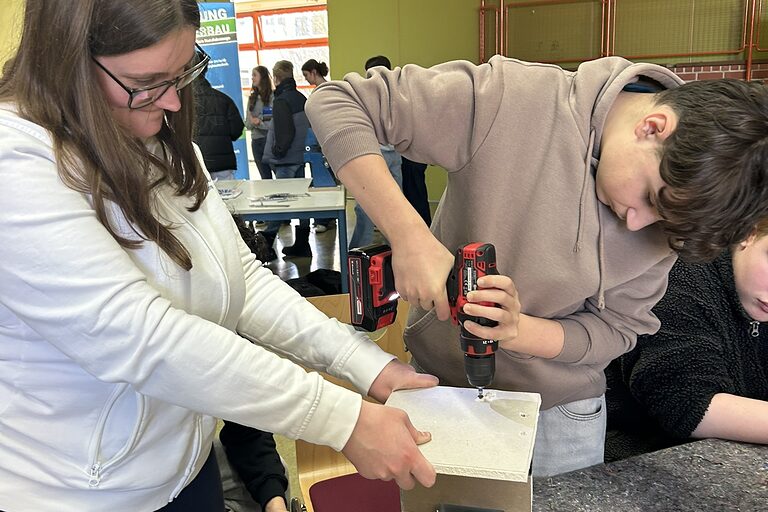 Ein Schüler dreht mit einem Akkuschrauber eine Schraube in ein Holzteil, das von einer Schülerin festgehalten wird.
