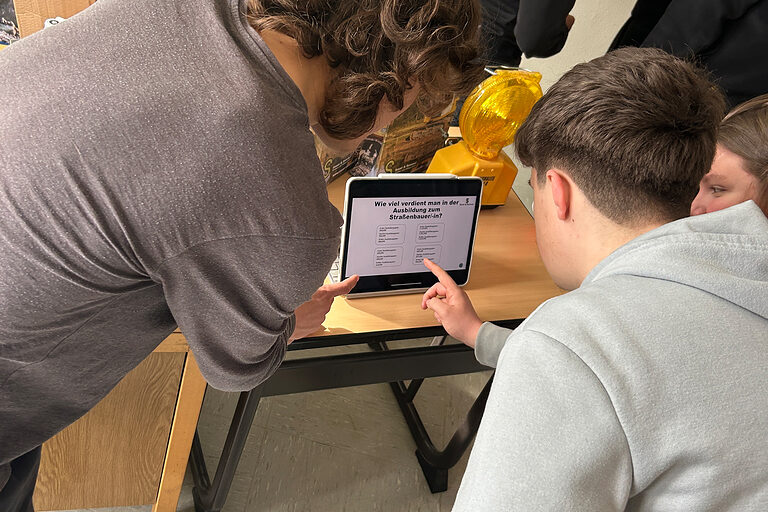Eine Schülergruppe beschäftigt sich mit einem Tablet an einem Stand eines Betriebs mit dem Beruf des Straßenbauers.
