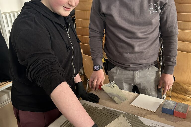 Eine Schülerin macht eine handwerkliche Übung. Ein Fliesen-, Platten- und Mosaikleger schaut zu.