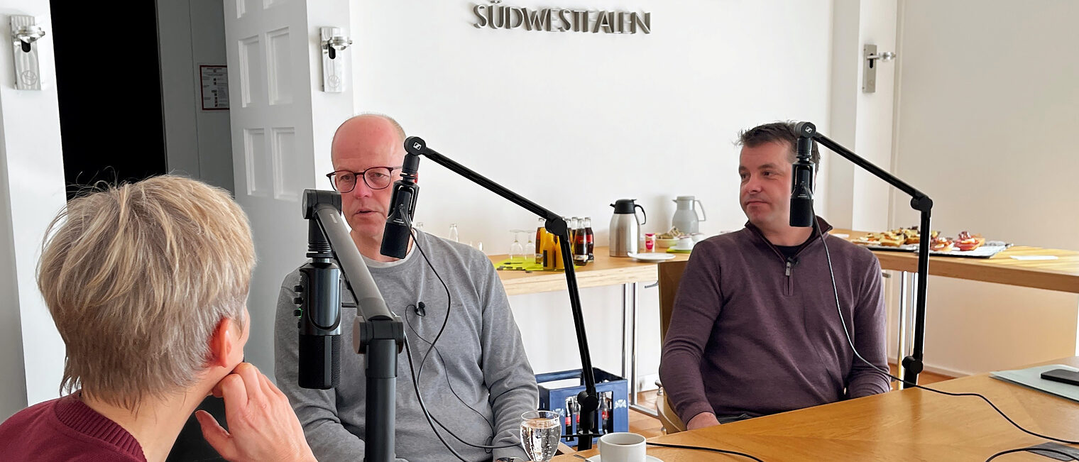 Eine Moderatorin und zwei Podcastgäste sitzen an einem Tisch. Jeder der drei hat ein Mikrofon vor sich.