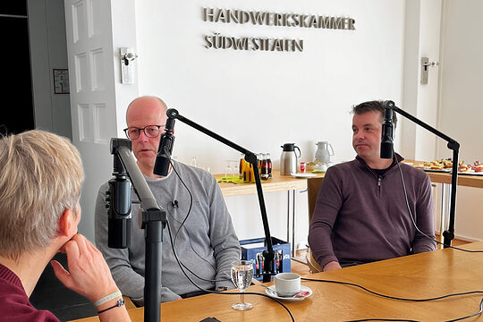 Eine Moderatorin und zwei Podcastgäste sitzen an einem Tisch. Jeder der drei hat ein Mikrofon vor sich.