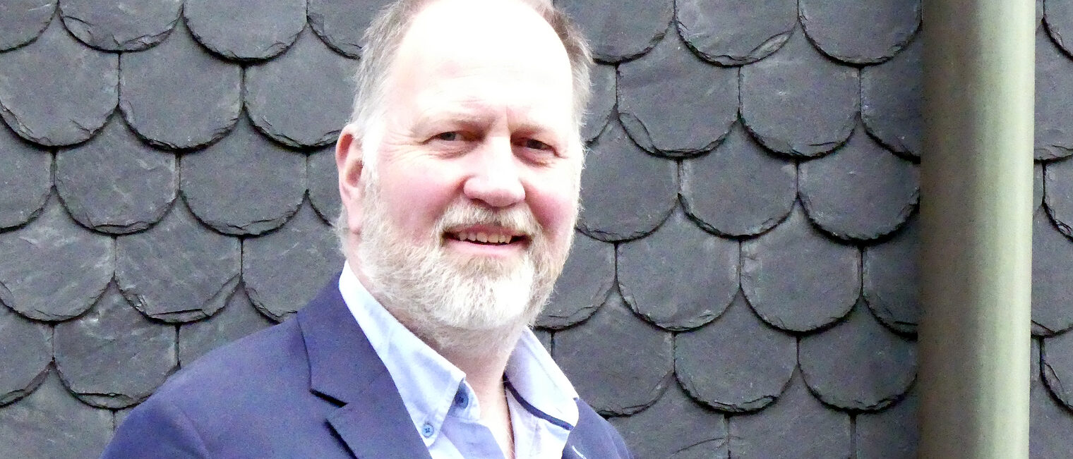 Porträt des Handwerkmeisters Frank Clemens. Er steht in einem Anzug vor einer Fassade in Schiefer-Optik.