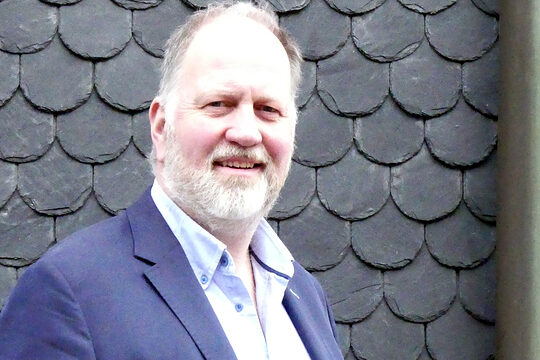 Porträt des Handwerkmeisters Frank Clemens. Er steht in einem Anzug vor einer Fassade in Schiefer-Optik.
