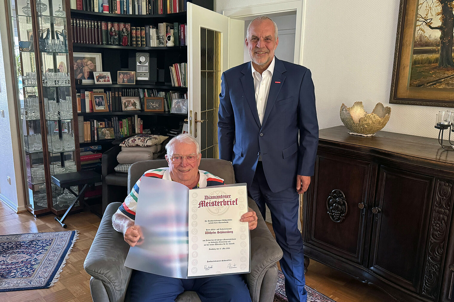 Wilhelm Schönenberg sitzt in einem Sessel und zeigt seinen Diamantenen Meisterbrief. HwK-Präsident Renfordt steht daneben.