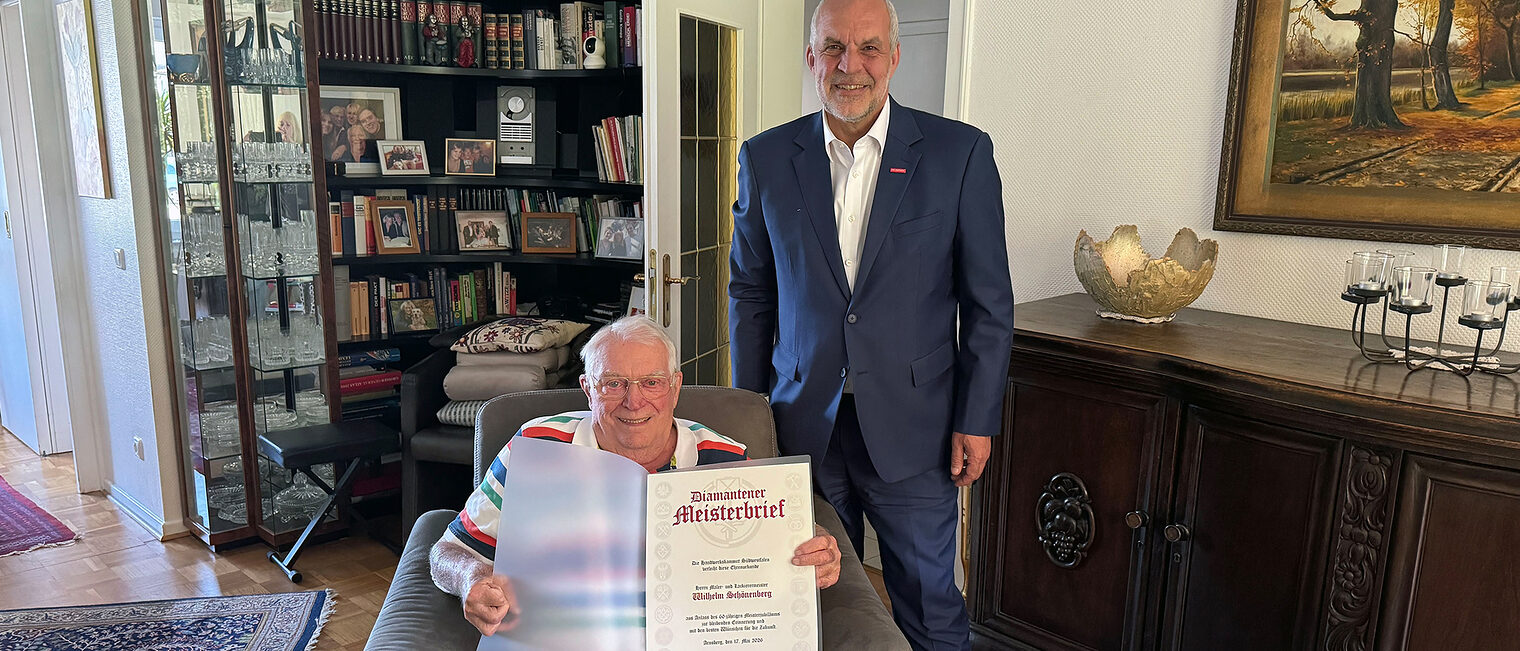 Wilhelm Schönenberg sitzt in einem Sessel und zeigt seinen Diamantenen Meisterbrief. HwK-Präsident Renfordt steht daneben.