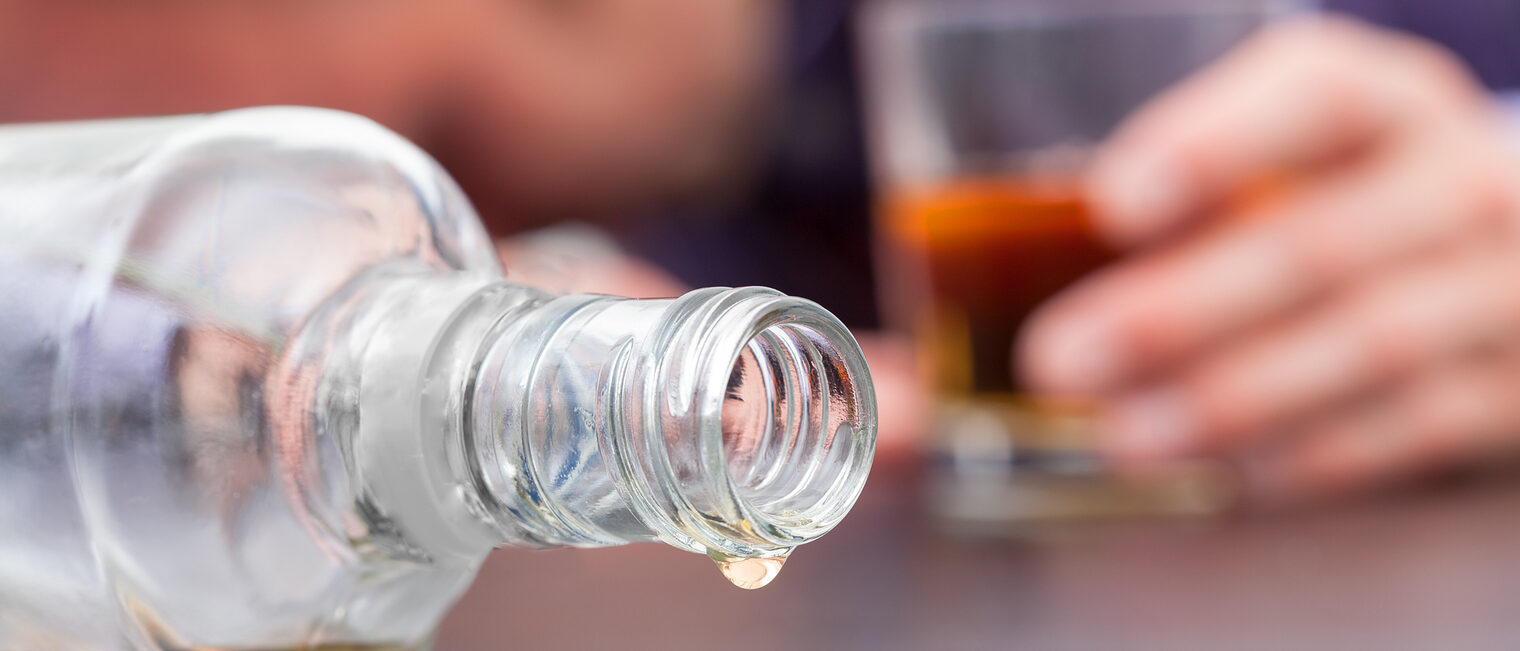 Eine umgekippte Glasflasche. Im Hintergrund ein Mann, der mit seinem Kopf auf dem Tisch liegt und ein Glas in der Hand hält.