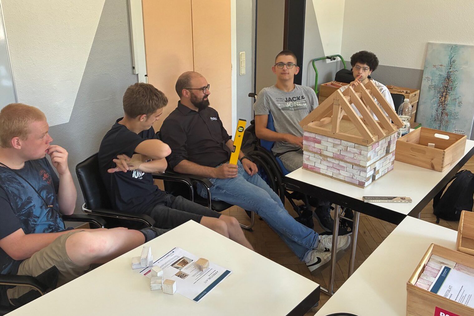 Ein Berater der Handwerkskammer Südwestfalen sitzt mit Workshop-Teilnehmenden an einem Tisch vor einem Modell eines Hauses.
