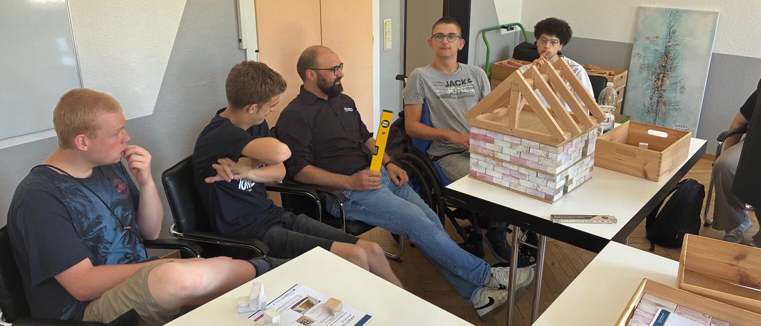 Ein Berater der Handwerkskammer Südwestfalen sitzt mit Workshop-Teilnehmenden an einem Tisch vor einem Modell eines Hauses.
