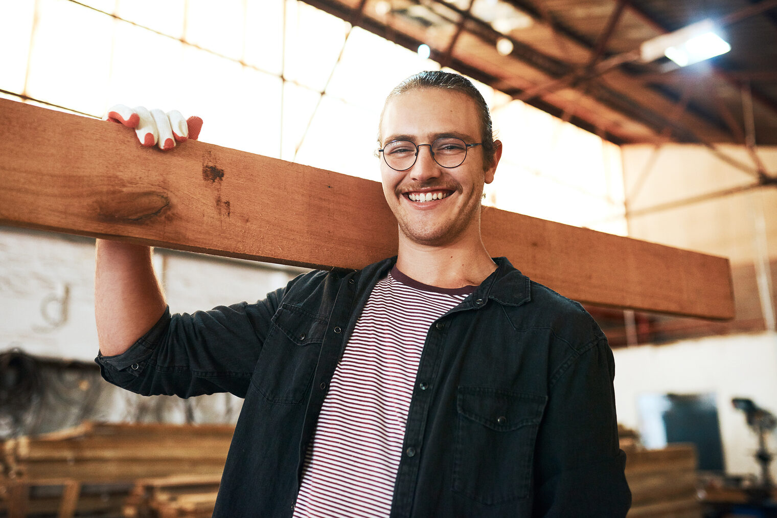 Portrait eines jungen Tischlers in seiner Werkstatt, der ein Holzbrett über die Schulter trägt.