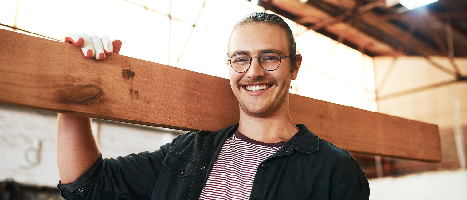 Portrait eines jungen Tischlers in seiner Werkstatt, der ein Holzbrett über die Schulter trägt.