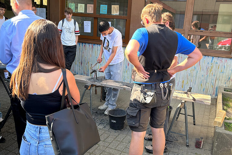 Ein Schüler schlägt aus Schiefer ein Herz aus, ein Dachdecker-Azubi und weitere Schüler sehen dabei zu.