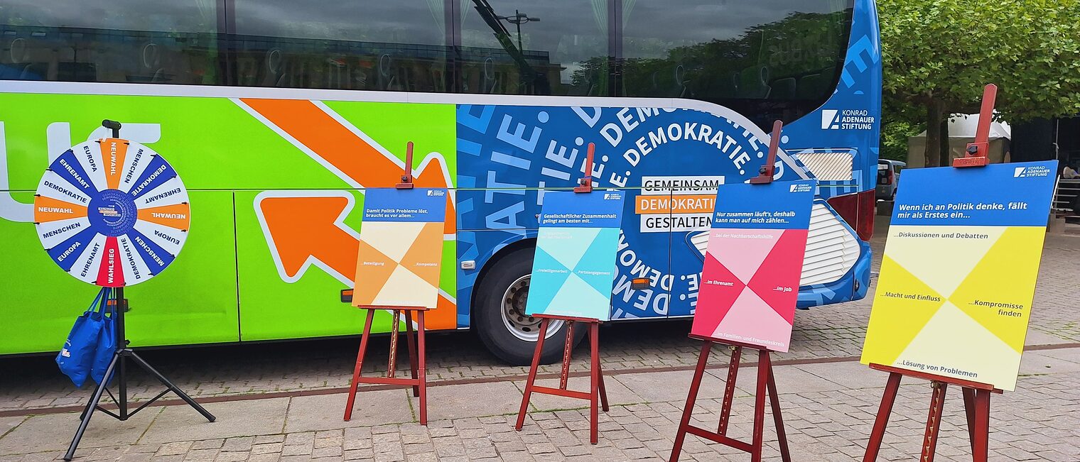 Ein Bus auf einem Parkplatz. Der Bus ist mit "Gemeinsam Demokratie gestalten" beschriftet. Vor dem Bus stehen Schaubilder.