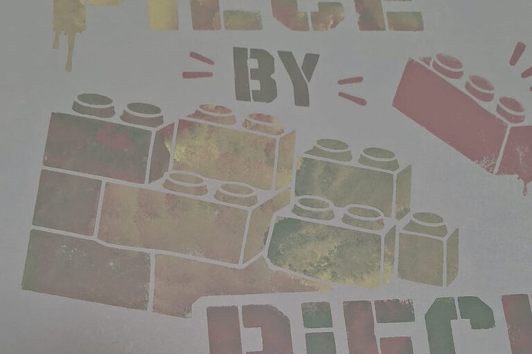 Gemalte Legosteine und der Schriftzug "Piece by Piece" auf einer hellen Platte.