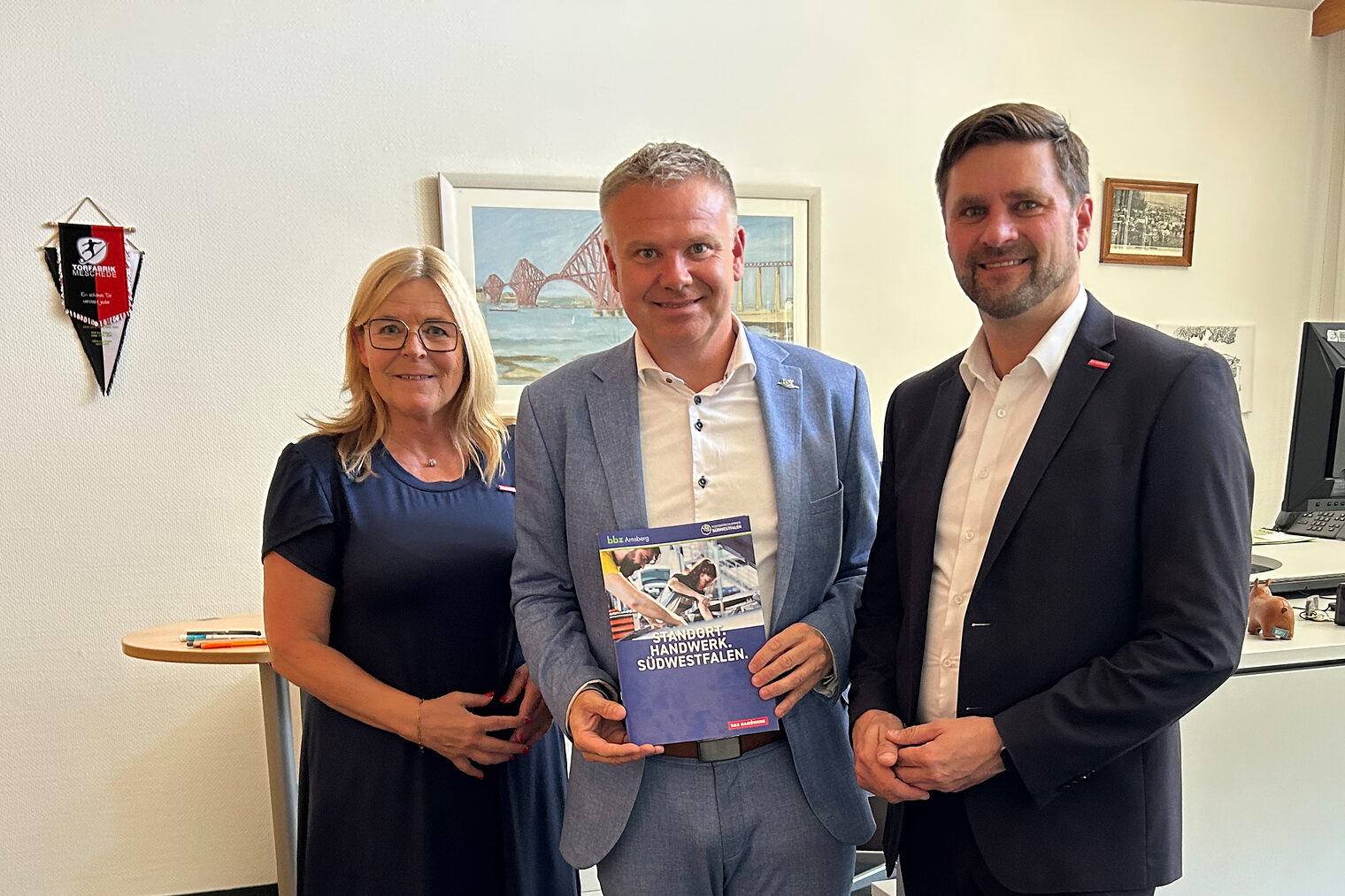 Gruppenbild mit Uta Neumeister, Thomas Grosche und Hendrik Schmitt. Grosche, in der Mitte stehend, hält eine Broschüre.
