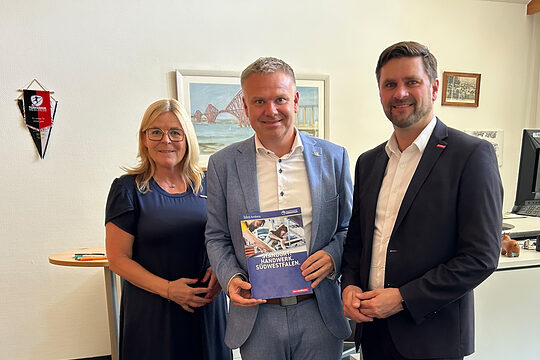 Gruppenbild mit Uta Neumeister, Thomas Grosche und Hendrik Schmitt. Grosche, in der Mitte stehend, hält eine Broschüre.