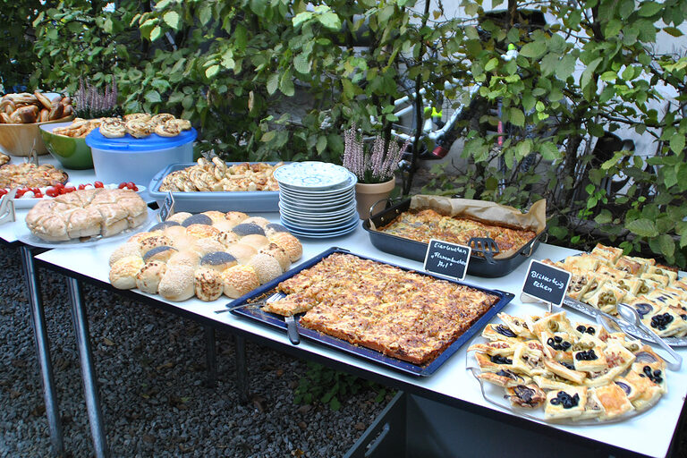 Verschiedene Ofenbleche mit Essen, zum Beispiel Zwiebelkuchen, verteilt auf zwei Tischen im Museumsgarten Haus Hövener.
