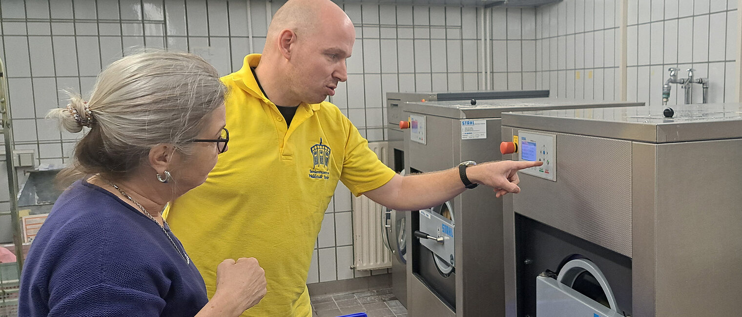 Ein gelb gekleideter Mitarbeiter erklärt einer Frau eine Waschmaschine in einer Wäscherei