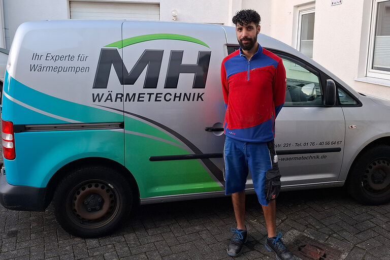 Ein junger Mann in Arbeitskleidung steht vor einem Firmenlieferwagen mit der Aufschrift "MH Wärmetechnik".