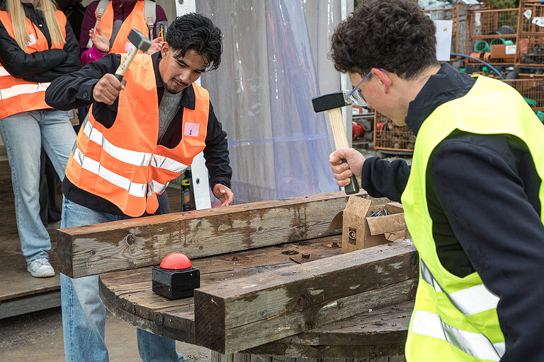 Zwei Schüler bearbeiten einen Holzbalken mit einem Hammer bei der Heckmann Bau Handwerkschallenge