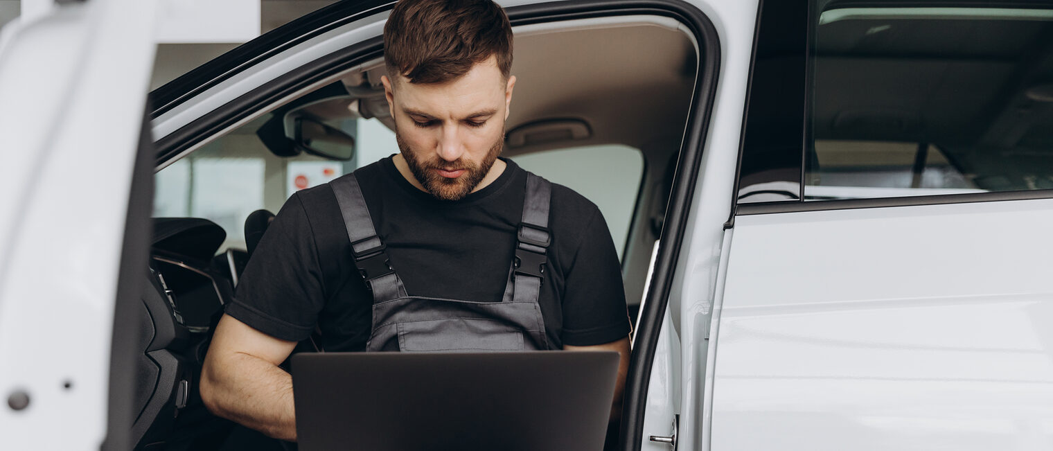Ein Kfz-Mechaniker sitzt in einem Fahrzeug mit geöffneter Tür und bedient ein Laptop.