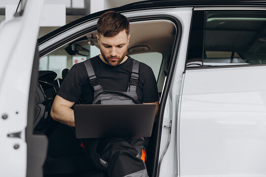 Ein Kfz-Mechaniker sitzt in einem Fahrzeug mit geöffneter Tür und bedient ein Laptop.