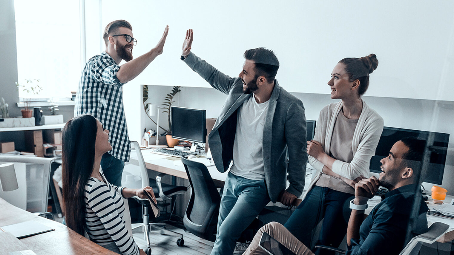 Two cheerful young business people giving high-five while their colleagues looking at them and smiling Schlagwort(e): Men, Women, People, Corporate Business, Modern, Success, Expertise, High-Five, Celebration, Group Of People, Office, Business, Creative Occupation, Multi-Ethnic Group, New Business, Solution, Working, Meeting, Brainstorming, Design Studio, Business Finance And Industry, Cooperation, Technology, Professional Occupation, Togetherness, Unity, Discussion, Smart Casual, Business Casual, Teamwork, Team, Ideas, Planning, Strategy, Coworker, Standing, Sitting, Leaning, Indoors, Talking, Smiling, Happiness, Gesturing, Beard, Transparent, Glass-Material