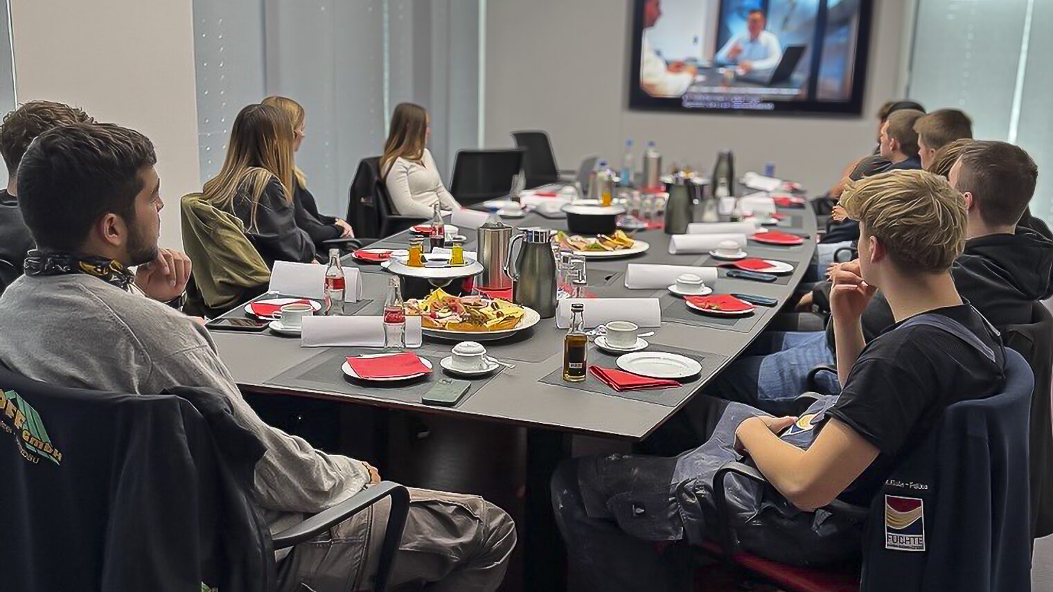 Zwölf Auszubildende sitzen in einem Schulungsraum an einem großen Tisch und schauen ein Schulungsvideo.